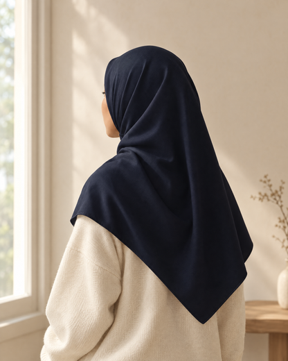 Perempuan berbaju rajut krem memakai bawal Biru Malam, terlihat dari belakang sambil menatap jendela
