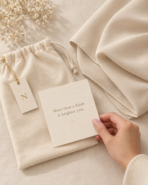 Pouch serut krem NURÉA, hang tag emas berinisial N, dan kartu ucapan bertuliskan More than a hijab. A brighter you.